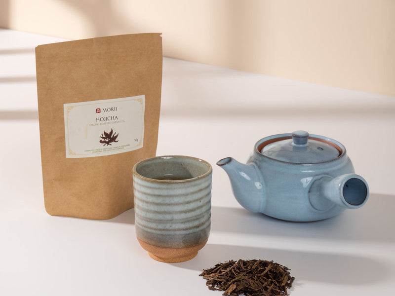 Morii hojicha pouch beside a teacup, blue teapot and loose roasted leaves