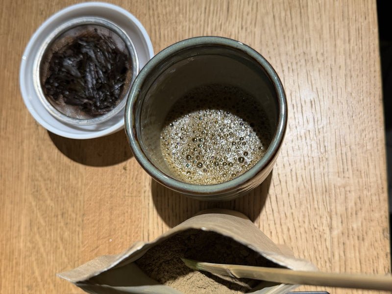 Open pack of Morii hojicha powder with a bamboo scoop on a scale, a cup of whisked hojicha and spent roasted leaves in a strainer, seen from above