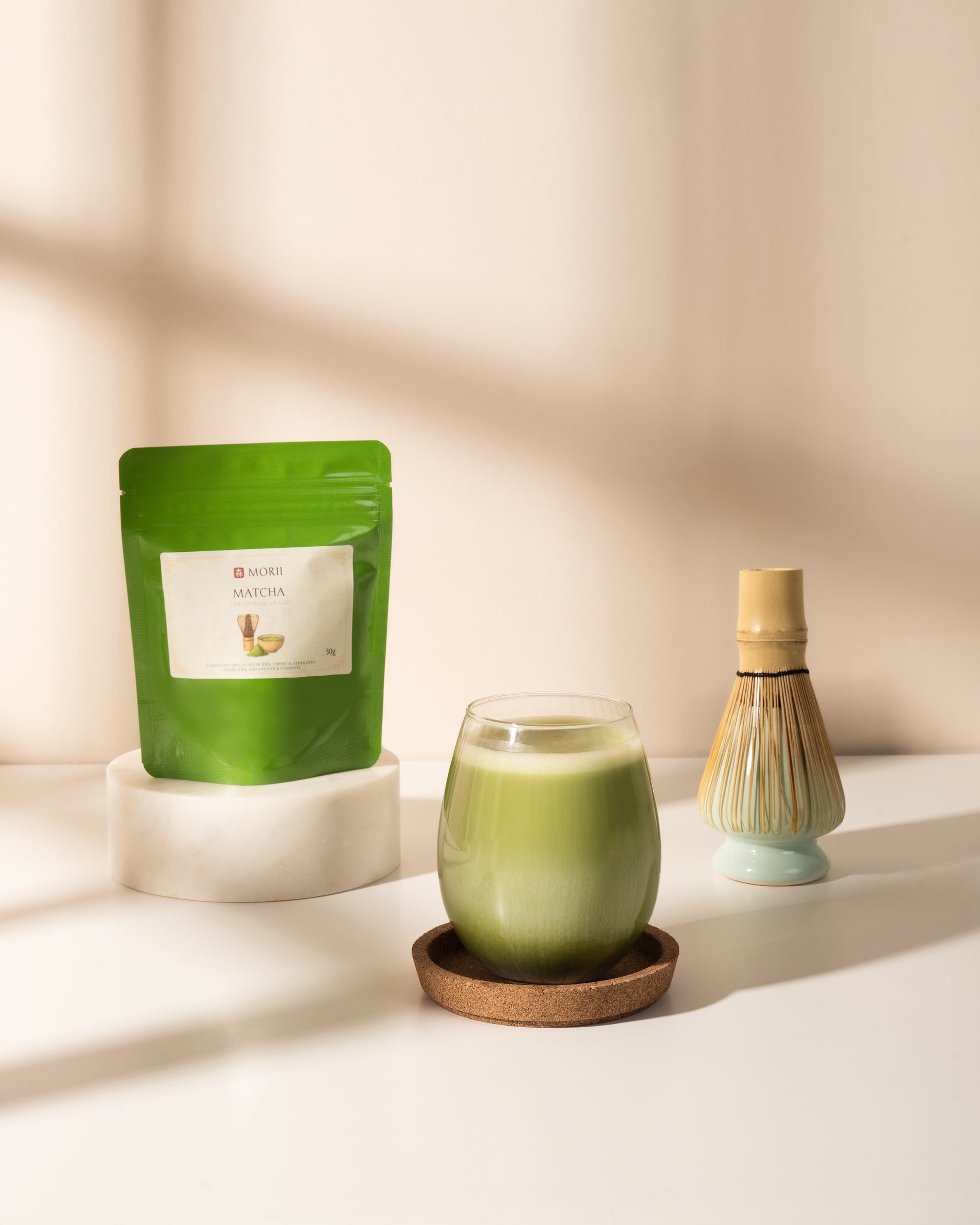 A green stand-up pouch labeled Matcha sits on a white pedestal beside a glass of light green matcha drink on a coaster and a bamboo whisk.